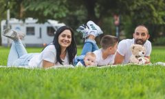 Happy family lying on grass with RV in the background, enjoying a sunny outdoor picnic.